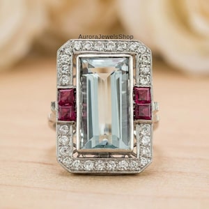 Pode incluir: Um anel retangular em estilo art déco com uma grande pedra central retangular azul clara. O anel é emoldurado com pequenos diamantes redondos e apresenta duas pedras vermelhas quadradas em cada lado. O anel é feito em metal prateado.