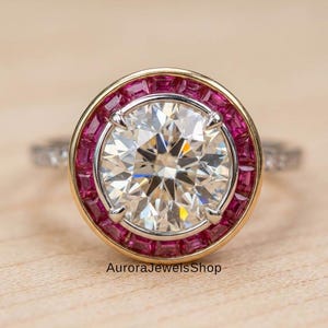 May include: A close-up of a diamond ring with a large, round, clear center stone. The ring has a gold band with a halo of small, square, pink gemstones. The text "AuroraJewelsShop" is at the bottom.