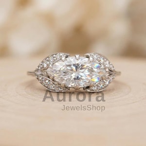 May include: A marquise-cut diamond ring with leaf-shaped detailing. The ring has a large, clear central stone and smaller diamonds set in the leaf designs. The text "Aurora JewelsShop" is visible.