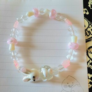 Op de afbeelding: Een kralenarmband met doorzichtige, roze en witte kralen. De armband bevat paddenstoelvormige kralen en een witte konijnenbedel. De armband ligt op gelijnd papier.