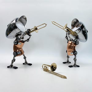 Puede incluir: Dos esculturas de robots tocando el trombón. Los robots tienen cabezas cromadas, torsos de cobre y piernas de metal. Un tercer trombón descansa sobre la superficie blanca. Las esculturas son artísticas y caprichosas.