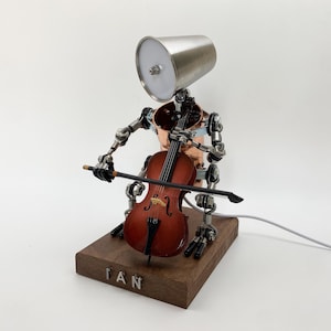 May include: A quirky robot sculpture playing a cello. The robot features a silver head, copper torso, and articulated metal limbs. The cello is a deep brown, and the sculpture is mounted on a wooden base with the name "IAN" engraved.