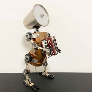 K&ouml;nnte beinhalten: Eine Roboter-Skulptur aus Silber und Kupfer, die ein rotes und schwarzes Akkordeon spielt. Der Roboter hat einen silbernen Lampenschirmkopf und steht auf einer schwarzen Oberfl&auml;che.