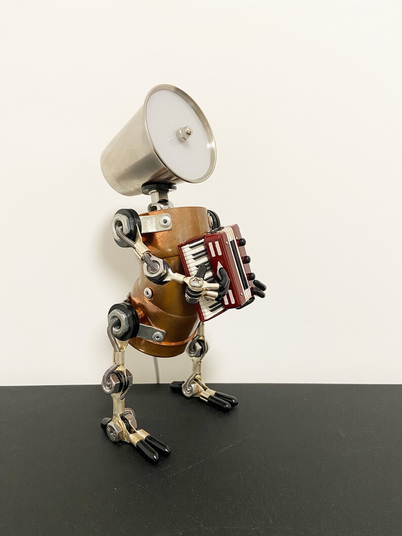 K&ouml;nnte beinhalten: Eine Roboter-Skulptur aus Silber und Kupfer, die ein rotes und schwarzes Akkordeon spielt. Der Roboter hat einen silbernen Lampenschirmkopf und steht auf einer schwarzen Oberfl&auml;che.
