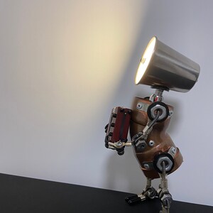 K&ouml;nnte beinhalten: Eine Steampunk-Roboterlampe aus Kupfer und Metallrohren. Der Roboter h&auml;lt eine kleine Holzkiste und hat einen silbernen Lampenschirm auf dem Kopf.