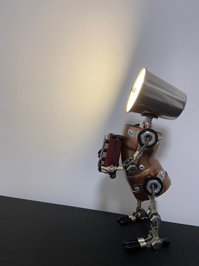K&ouml;nnte beinhalten: Eine Steampunk-Roboterlampe aus Kupfer und Metallrohren. Der Roboter h&auml;lt eine kleine Holzkiste und hat einen silbernen Lampenschirm auf dem Kopf.