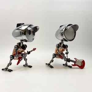 May include: Two robot sculptures, each with a chrome headlight for a head, copper torso, and metal legs. One robot wears sunglasses and plays a red guitar. The other robot plays a red guitar. The sculptures are on a white background.