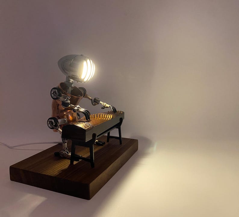 May include: A steampunk-style lamp shaped like a robot playing a zither. The robot is made of copper and metal, and the zither is made of wood. The lamp is on a wooden base.
