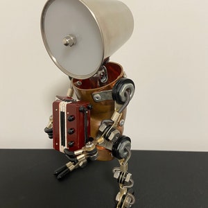 K&ouml;nnte beinhalten: Ein kleiner, skurriler Roboter aus Metall und Kupfer, mit einem silbernen Lampenschirm als Kopf, der ein rotes Akkordeon spielt. Der Roboter hat bewegliche Beine und Arme und sitzt auf einer schwarzen Oberfl&auml;che.