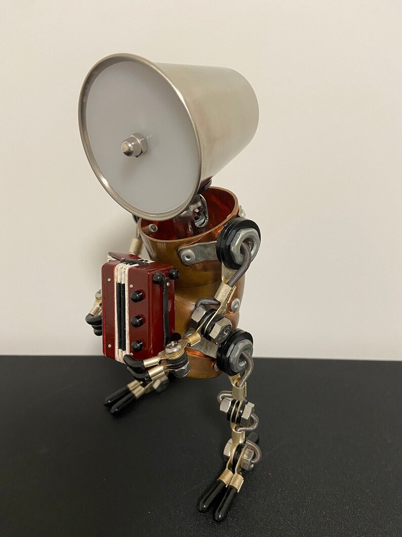 K&ouml;nnte beinhalten: Ein kleiner, skurriler Roboter aus Metall und Kupfer, mit einem silbernen Lampenschirm als Kopf, der ein rotes Akkordeon spielt. Der Roboter hat bewegliche Beine und Arme und sitzt auf einer schwarzen Oberfl&auml;che.