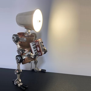 K&ouml;nnte beinhalten: Eine Steampunk-Lampe aus wiederverwendeten Materialien mit einem Kupferk&ouml;rper, einem silbernen Lampenschirm und einem kleinen Akkordeon. Die Lampe ist eingeschaltet und strahlt ein warmes Licht aus.