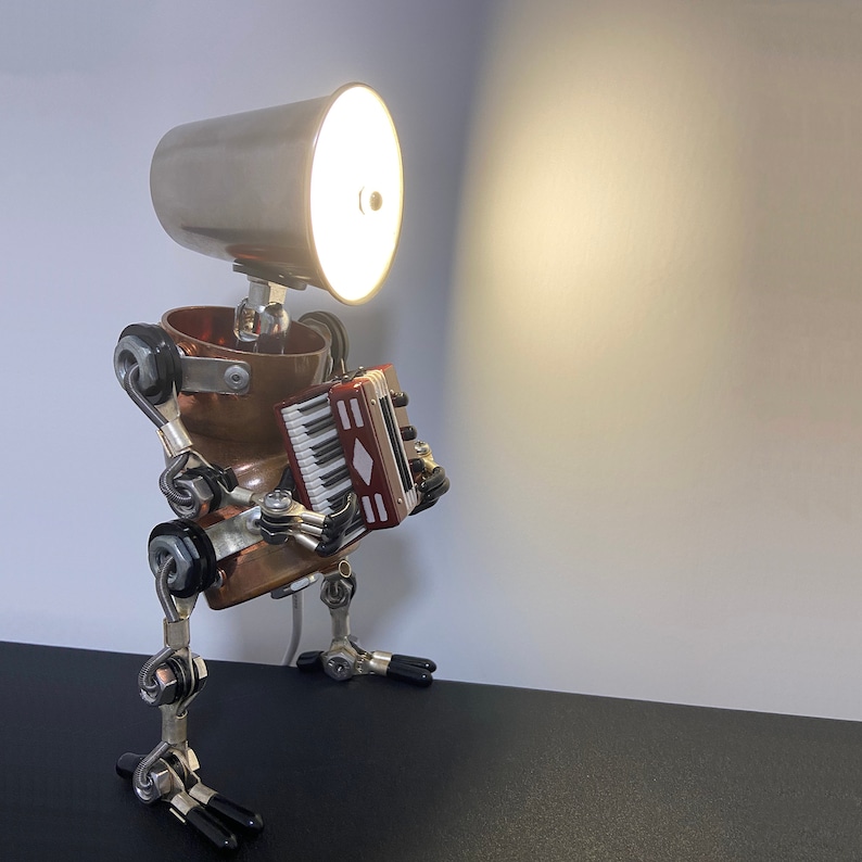 K&ouml;nnte beinhalten: Eine Steampunk-Lampe aus wiederverwendeten Materialien mit einem Kupferk&ouml;rper, einem silbernen Lampenschirm und einem kleinen Akkordeon. Die Lampe ist eingeschaltet und strahlt ein warmes Licht aus.