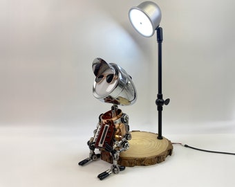 May include: A silver and copper robot with sunglasses and a hat sits on a wooden base. A black lamp with a silver shade is positioned behind the robot.