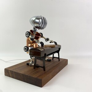 May include: A robot made of metal and copper plays a zither, a traditional Chinese musical instrument. The robot is sitting on a wooden base.