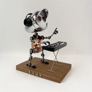 May include: A quirky robot sculpture crafted from metal parts, playing a miniature keyboard. The robot features a silver head with sunglasses, a copper torso, and articulated limbs. It rests on a wooden base with the word "love" spelled out.