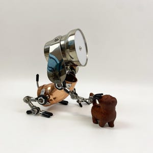 Può includere: Un'esclusiva scultura di cane in metallo realizzata a mano, con corpo in rame, testa cromata e arti articolati. Il cane interagisce con una piccola statuina di cane in legno marrone. La scultura ha un'estetica meccanica e industriale.