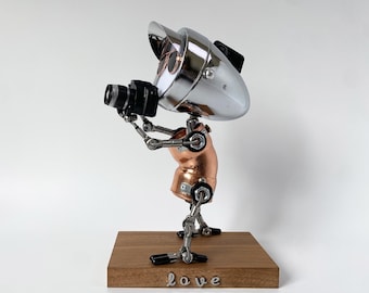 May include: A metal sculpture of a figure holding a camera, featuring a chrome head with a visor and sunglasses, a copper-colored torso, and articulated limbs. The figure stands on a rectangular wooden base with the word love in metal letters.