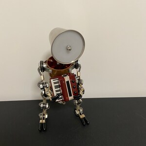 K&ouml;nnte beinhalten: Ein silberner Roboter mit einem Lampenschirmkopf und einem roten und schwarzen Akkordeon. Der Roboter hat bewegliche Beine und Arme.