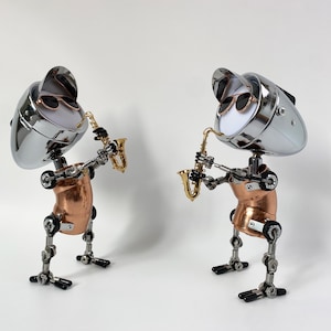 Puede incluir: Dos esculturas de robots, cada una tocando un saxofón dorado. Los robots tienen cabezas cromadas con gafas de sol, torsos de cobre y extremidades metálicas articuladas. Se encuentran sobre una superficie blanca.