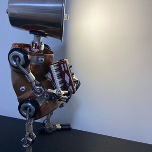K&ouml;nnte beinhalten: Ein Roboter im Steampunk-Stil aus Metallrohren und Zahnr&auml;dern, der ein kleines rotes Akkordeon h&auml;lt. Der Roboter hat einen silbernen Lampenschirm als Kopf und steht auf einer schwarzen Oberfl&auml;che.
