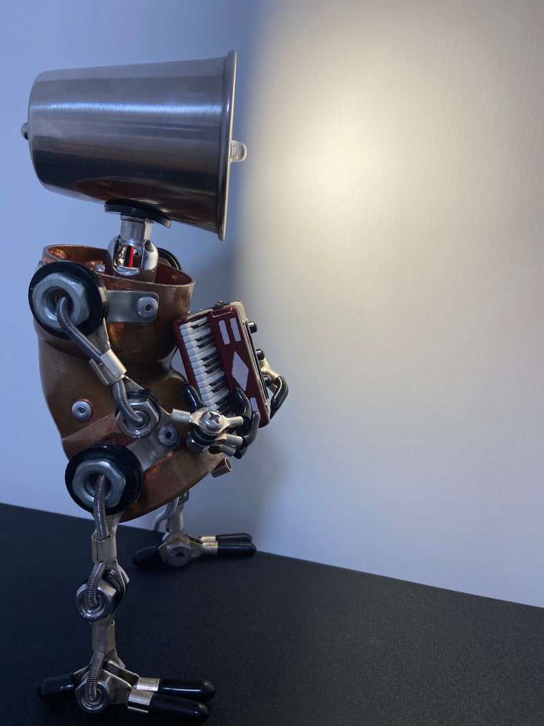 K&ouml;nnte beinhalten: Ein Roboter im Steampunk-Stil aus Metallrohren und Zahnr&auml;dern, der ein kleines rotes Akkordeon h&auml;lt. Der Roboter hat einen silbernen Lampenschirm als Kopf und steht auf einer schwarzen Oberfl&auml;che.