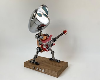 Puede incluir: Escultura metálica única de un robot tocando una guitarra eléctrica roja. El robot tiene una cabeza cromada con gafas de sol, un torso de cobre y piernas articuladas. Se encuentra sobre una base de madera con la palabra "Abod" grabada.