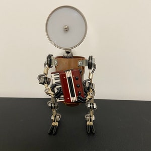 K&ouml;nnte beinhalten: Eine kleine Metallsculptur eines Roboters, der Akkordeon spielt. Der Roboter besteht aus Kupfer- und Silbermetallteilen und hat einen runden, silbernen Kopf mit einem wei&szlig;en Licht. Der Roboter h&auml;lt ein rotes und schwarzes Akkordeon mit silbernen Tasten.