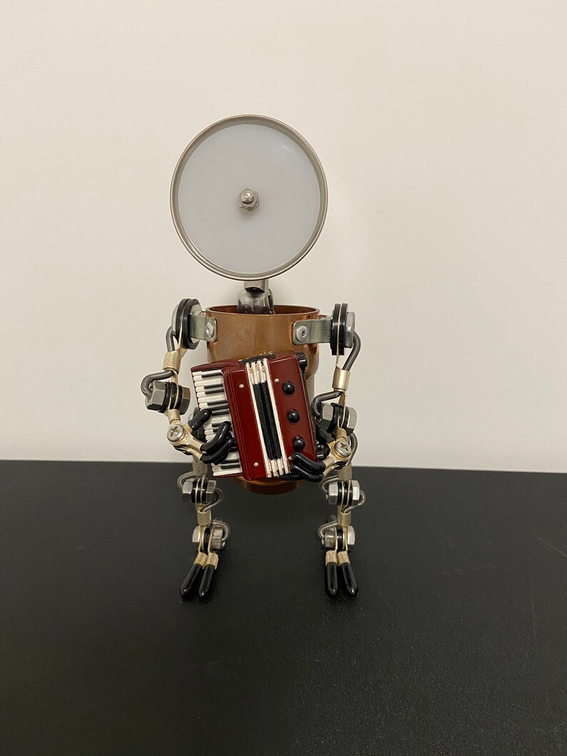 K&ouml;nnte beinhalten: Eine kleine Metallsculptur eines Roboters, der Akkordeon spielt. Der Roboter besteht aus Kupfer- und Silbermetallteilen und hat einen runden, silbernen Kopf mit einem wei&szlig;en Licht. Der Roboter h&auml;lt ein rotes und schwarzes Akkordeon mit silbernen Tasten.