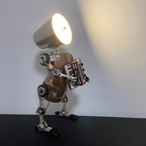 K&ouml;nnte beinhalten: Eine silberne und kupferne Roboterlampe mit eingeschaltetem Licht, die ein rotes Akkordeon spielt. Der Roboter hat einen runden Kopf mit einem silbernen Lampenschirm und einen kupfernen K&ouml;rper. Der Roboter hat bewegliche Arme und Beine.