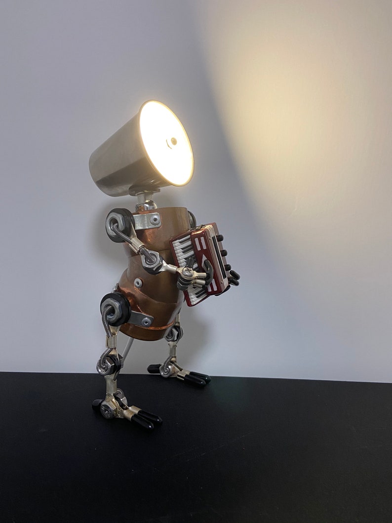 K&ouml;nnte beinhalten: Eine silberne und kupferne Roboterlampe mit eingeschaltetem Licht, die ein rotes Akkordeon spielt. Der Roboter hat einen runden Kopf mit einem silbernen Lampenschirm und einen kupfernen K&ouml;rper. Der Roboter hat bewegliche Arme und Beine.