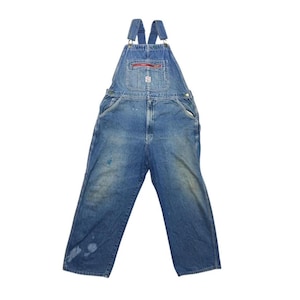 Könnte beinhalten: Blaue Jeans-Latzhose mit verstellbaren Trägern und einer Fronttasche. Die Latzhose hat einen roten Reißverschluss an der Brusttasche und mehrere Taschen. Der Denim hat ein verwaschenes, abgenutztes Aussehen.