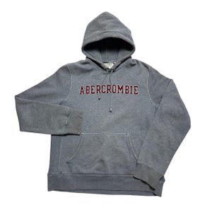 May include: A gray Abercrombie hoodie with a drawstring hood and a kangaroo pocket. The word "Abercrombie" is embroidered across the chest in maroon lettering with white trim. The sweatshirt is made of a soft, comfortable material.