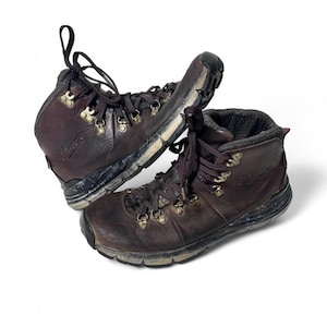 May include: A pair of worn brown leather hiking boots with dark brown laces and gold-colored eyelets. The boots show signs of wear and tear, with scuff marks and dirt visible on the soles and uppers.