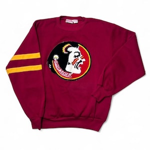 May include: A maroon long-sleeve sweatshirt featuring two horizontal yellow stripes on the sleeves and a circular patch on the chest. The patch displays a white profile of a person with black hair and the text Florida State in white lettering.