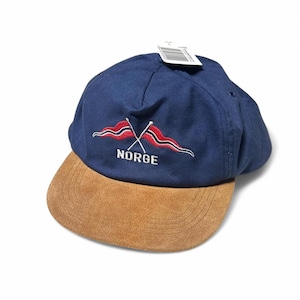 以下が含まれることがあります： スエードのつばが付いた青と茶色のベースボールキャップ。キャップの前方には、2本の交差した旗と「NORGE」という文字が白で刺繍されています。