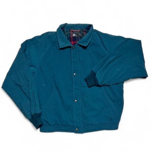 May include: Teal-colored jacket with a collared neckline and button-up front. The jacket features a dark green ribbed cuff and a red and green plaid lining visible at the collar. The jacket has two front pockets and a ribbed hem. A classic outerwear piece.