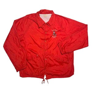 May include: A bright red vintage coach jacket with a button-up front, collar, and drawstring hem. The jacket features a small embroidered design on the chest with the text "M.C. State" and a cartoon character. The jacket is made of a lightweight, possibly nylon, material.