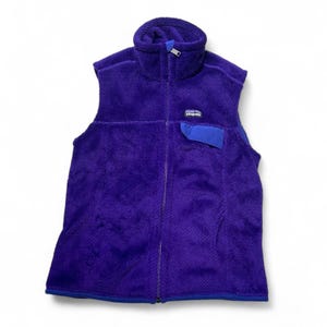 May include: A sleeveless, purple fleece vest with a high collar and a full-length zipper. The vest features a blue pocket on the chest and a small logo patch. The bottom hem and pocket are trimmed with blue fabric.