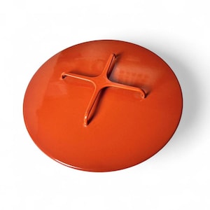 May include: A circular, glossy orange lid featuring a centered, raised cross-shaped handle. The lid has a smooth, solid-colored finish and a simple, functional design, providing a secure cover for a matching container or pot.