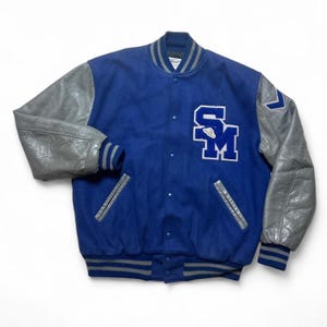 May include: A blue and gray varsity jacket with gray leather sleeves. The jacket features a button-up front, striped cuffs, and a large "SM" patch on the chest. The jacket has two front pockets and a striped collar.