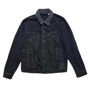 May include: A black denim jacket with a gray sherpa collar. The jacket has two chest pockets, two side pockets, and a button-up front. A small red tag is visible on the chest pocket. The jacket is a classic style.