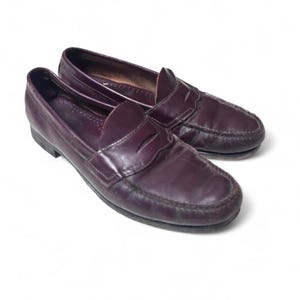 May include: A pair of burgundy leather penny loafers. The shoes have a classic design with a rounded toe, a strap across the vamp, and a low heel. The leather has a slight sheen, and the shoes appear to be well-worn.