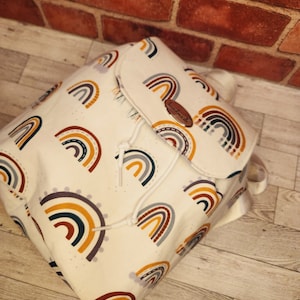 May include: A white canvas backpack with a drawstring closure. The backpack is decorated with a colorful rainbow pattern and the word "handmade" is printed on a brown leather tag.