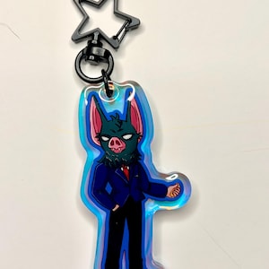 May include: A keychain featuring a cartoon bat character in a suit, with a star-shaped clasp. The bat has a green face, pink ears, and is wearing a blue suit, black pants, and a red tie. The keychain has an iridescent border.