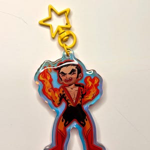 May include: A colorful acrylic keychain featuring a cartoon character with flames emanating from their hands. The character is wearing a black and red outfit. The keychain has a yellow star-shaped clasp.