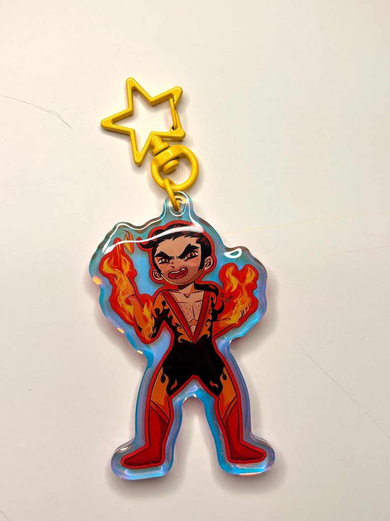 May include: A colorful acrylic keychain featuring a cartoon character with flames emanating from their hands. The character is wearing a black and red outfit. The keychain has a yellow star-shaped clasp.