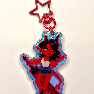 May include: A red devil-themed keychain with a star-shaped clasp. The keychain features a cartoon illustration of a red-skinned character with black hair, horns, and a tail, wearing a white tank top, denim shorts, and black heels. The character is holding a black object.
