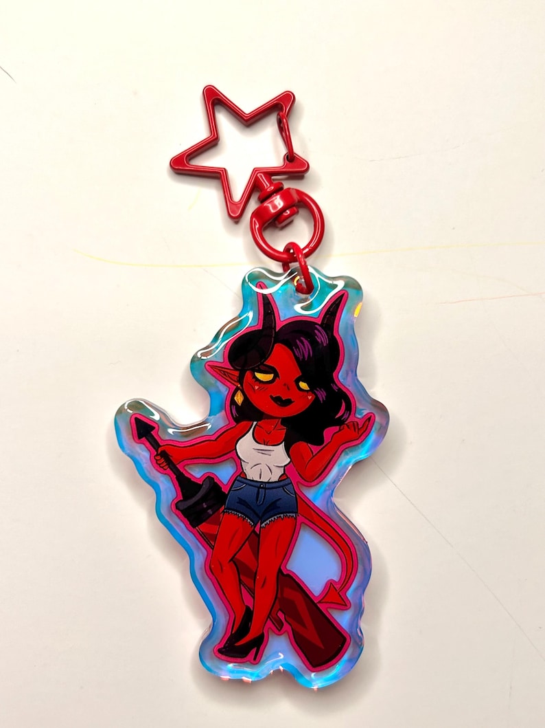 May include: A red devil-themed keychain with a star-shaped clasp. The keychain features a cartoon illustration of a red-skinned character with black hair, horns, and a tail, wearing a white tank top, denim shorts, and black heels. The character is holding a black object.