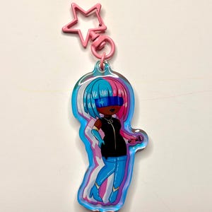 May include: A colorful keychain featuring a cartoon character with blue and pink hair, wearing a black outfit and blue boots. The keychain has a pink star-shaped clasp and a silver ring. The character is holding a microphone.