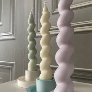 May include: Three spiral candles in different colours, green, white, and purple, on white ceramic candle holders.